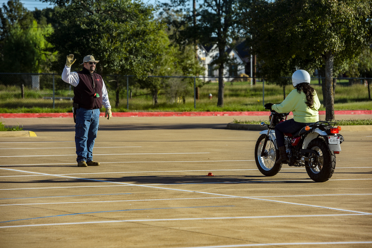 Standing in an open parking lot, Motorcycle riding instructor waves his hand to direct his student motorcycle rider.